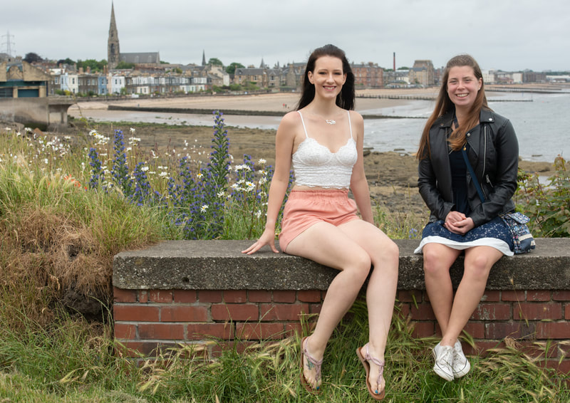 Two young women sit on a wall with the houses on Portobello promenade behind them