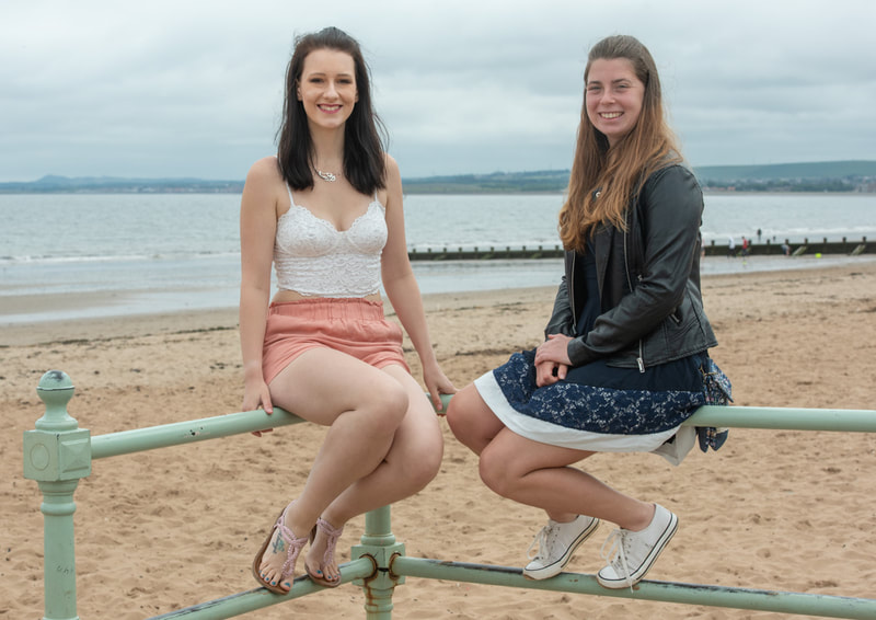Two young women sit on the prom railings with the beach behind them