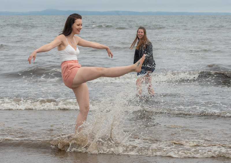 A young woman in the foreground kicks up the water while her friend, a little further out into the sea looks on.